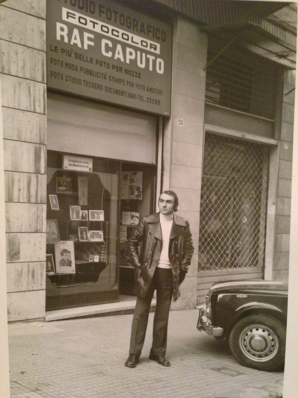 Vintage black and white photo of a storefront with RAF photo sign and parked car from the 1950s.