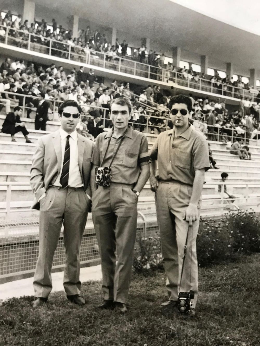 Group standing on grass field in front of stadium seating wearing suits in vintage photograph.