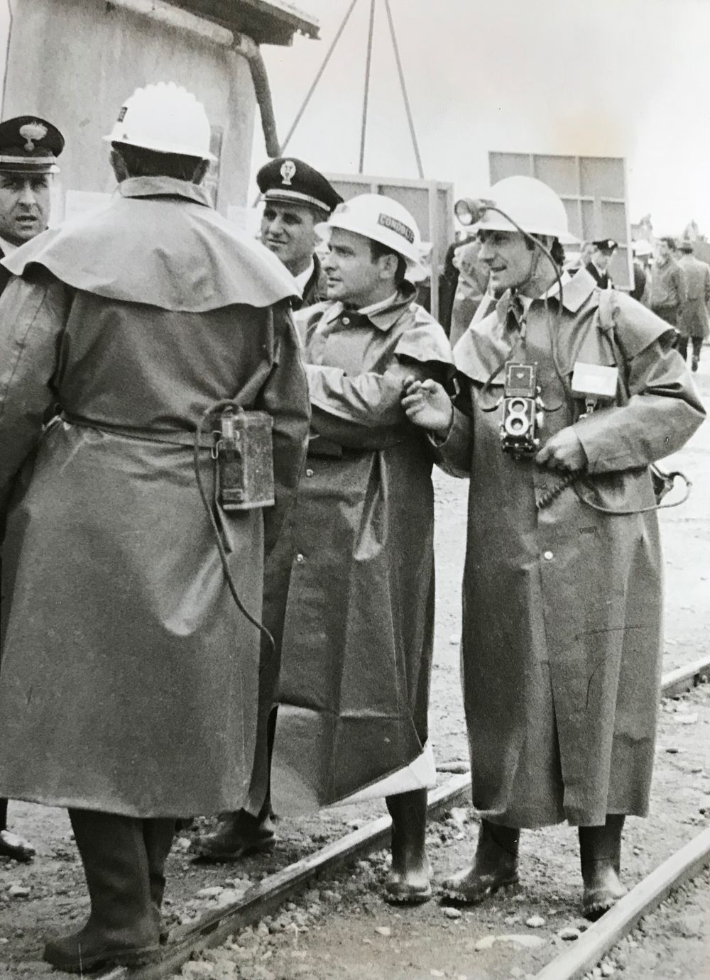 Group of people in coats gathered around equipment outdoors in black and white photograph.
