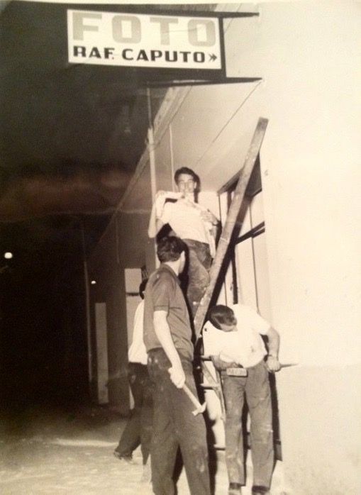Vintage black and white photograph showing people standing near a storefront sign.