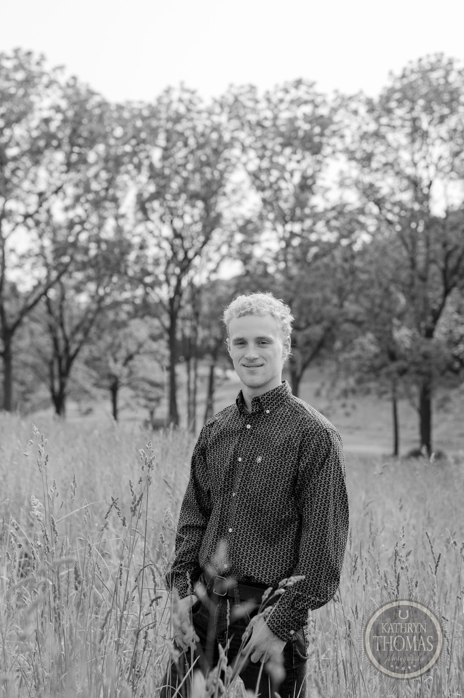 Black and white portrait in field of tall grass with grove of trees in background.