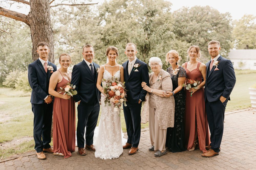 Wedding party group photo with bride, groom, bridesmaids in rust gowns and groomsmen in navy suits.