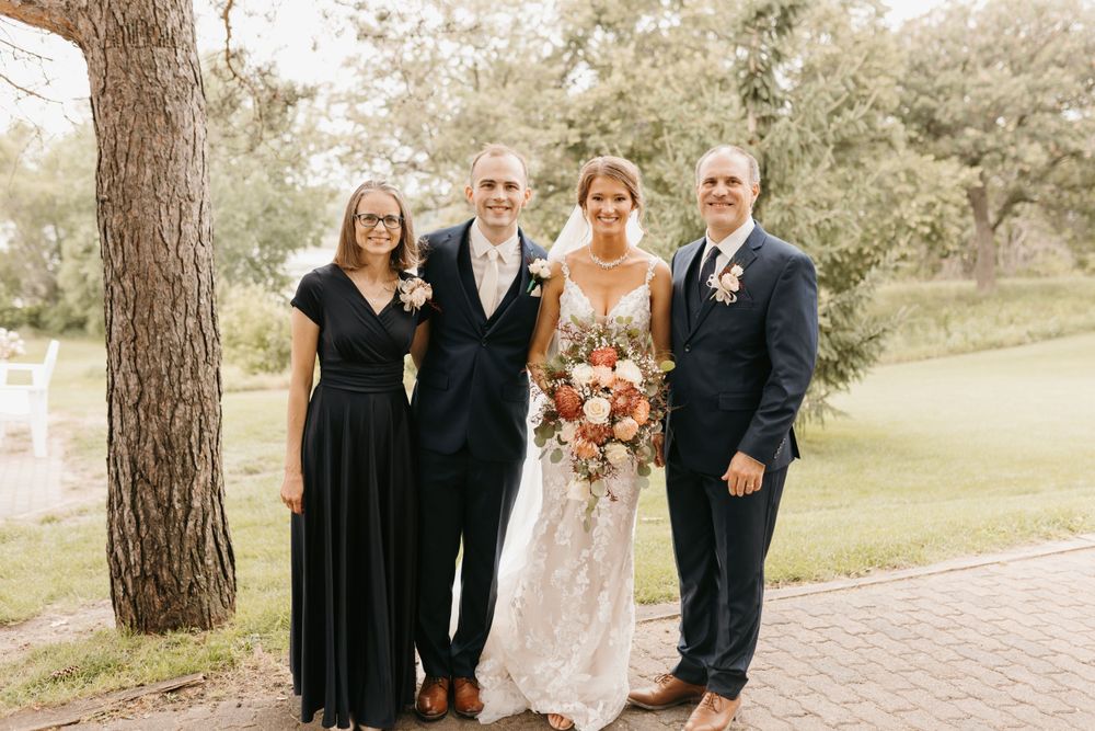Bride holding floral bouquet poses with groom and two family members in black attire outdoors.