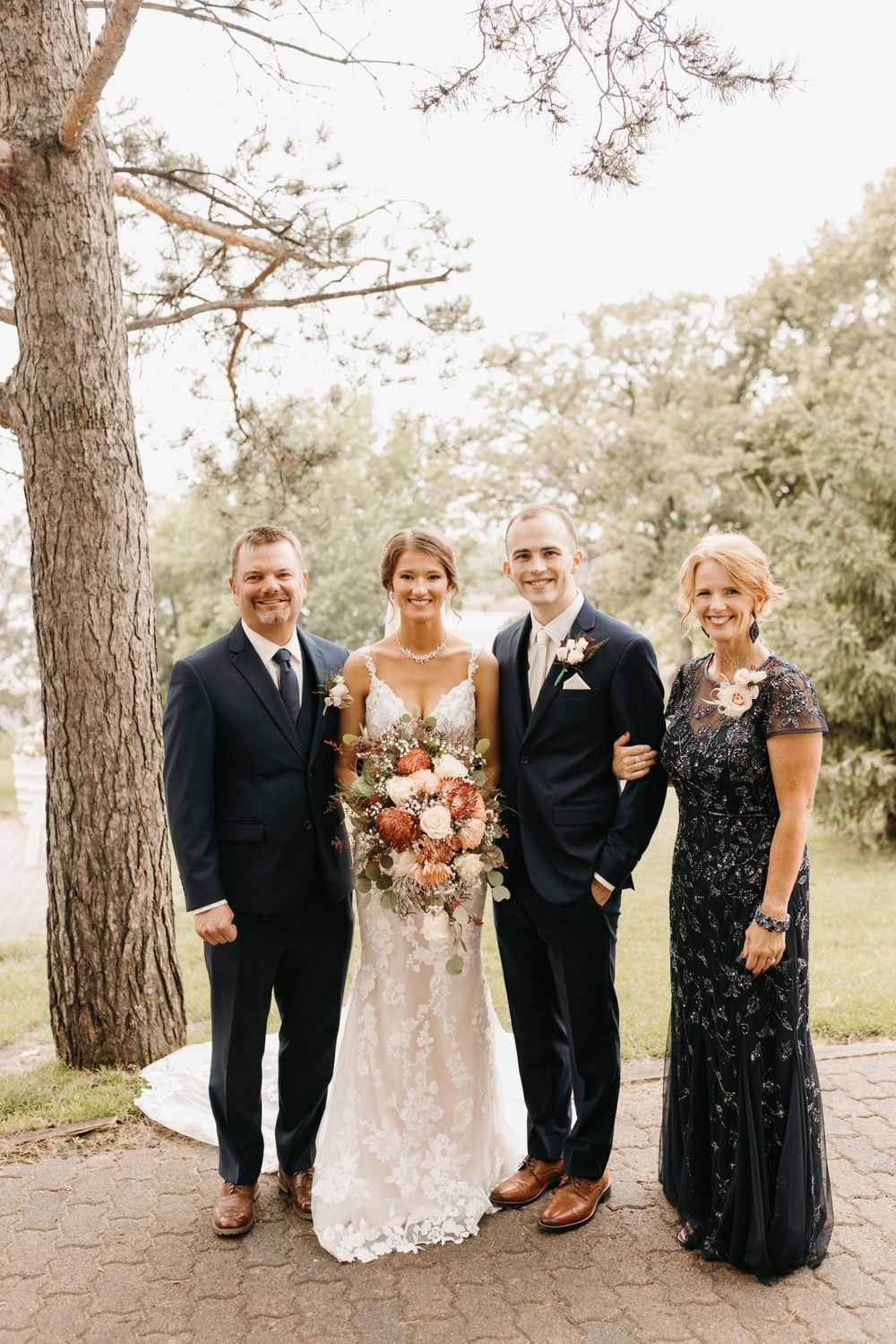 Bride and groom pose with two family members outdoors at a wedding venue surrounded by trees.