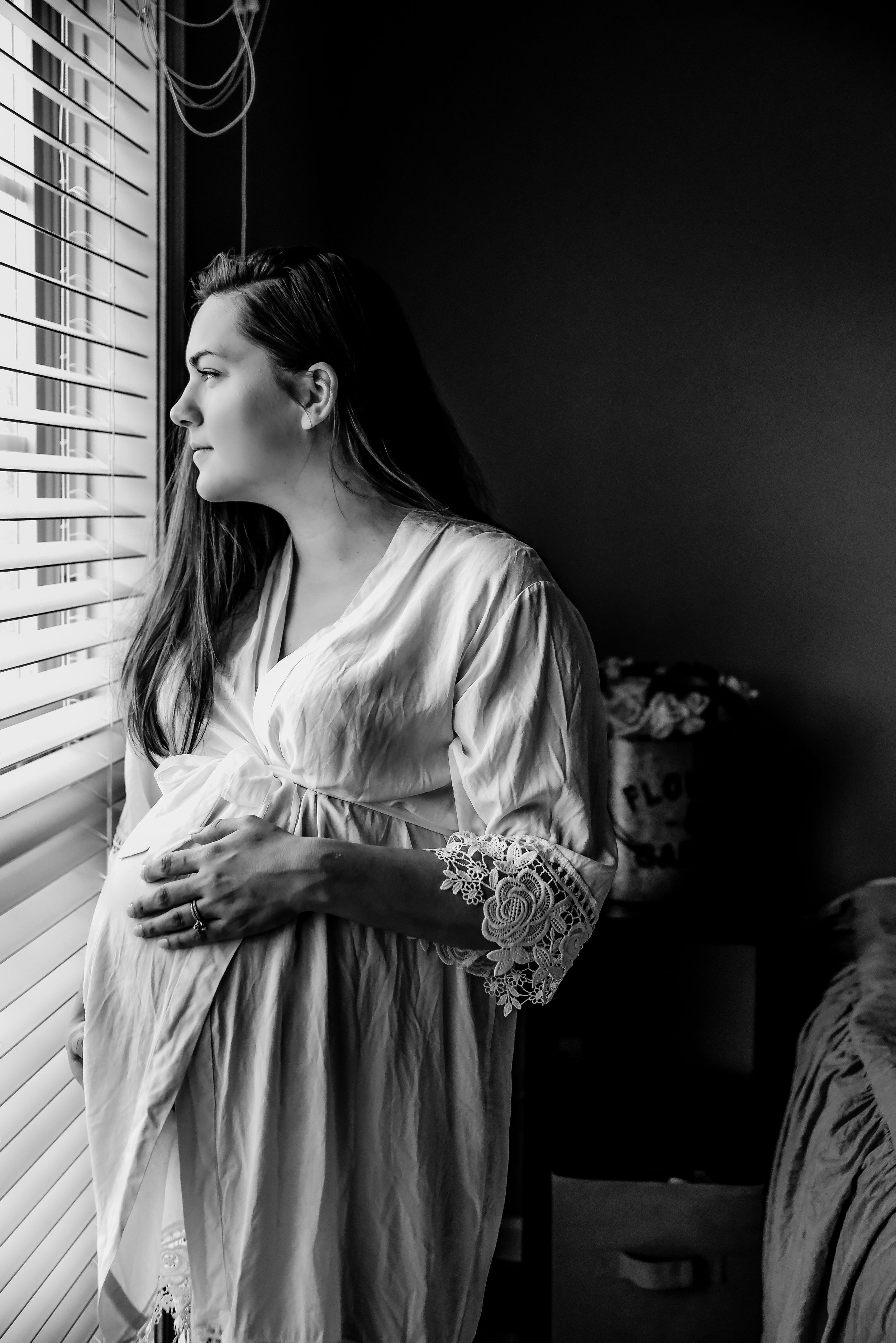 Artistic black and white portrait of woman in white dress by window with blinds.