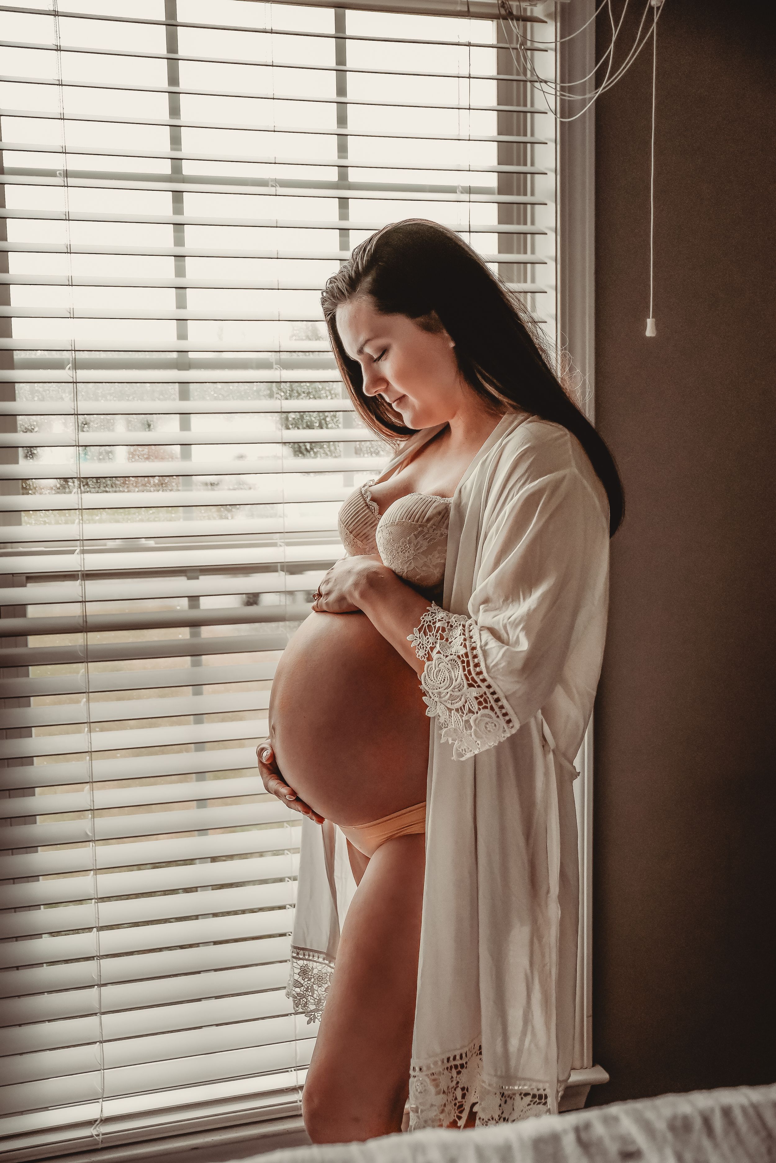 Side view of pregnant woman in white dress standing by window with natural light.