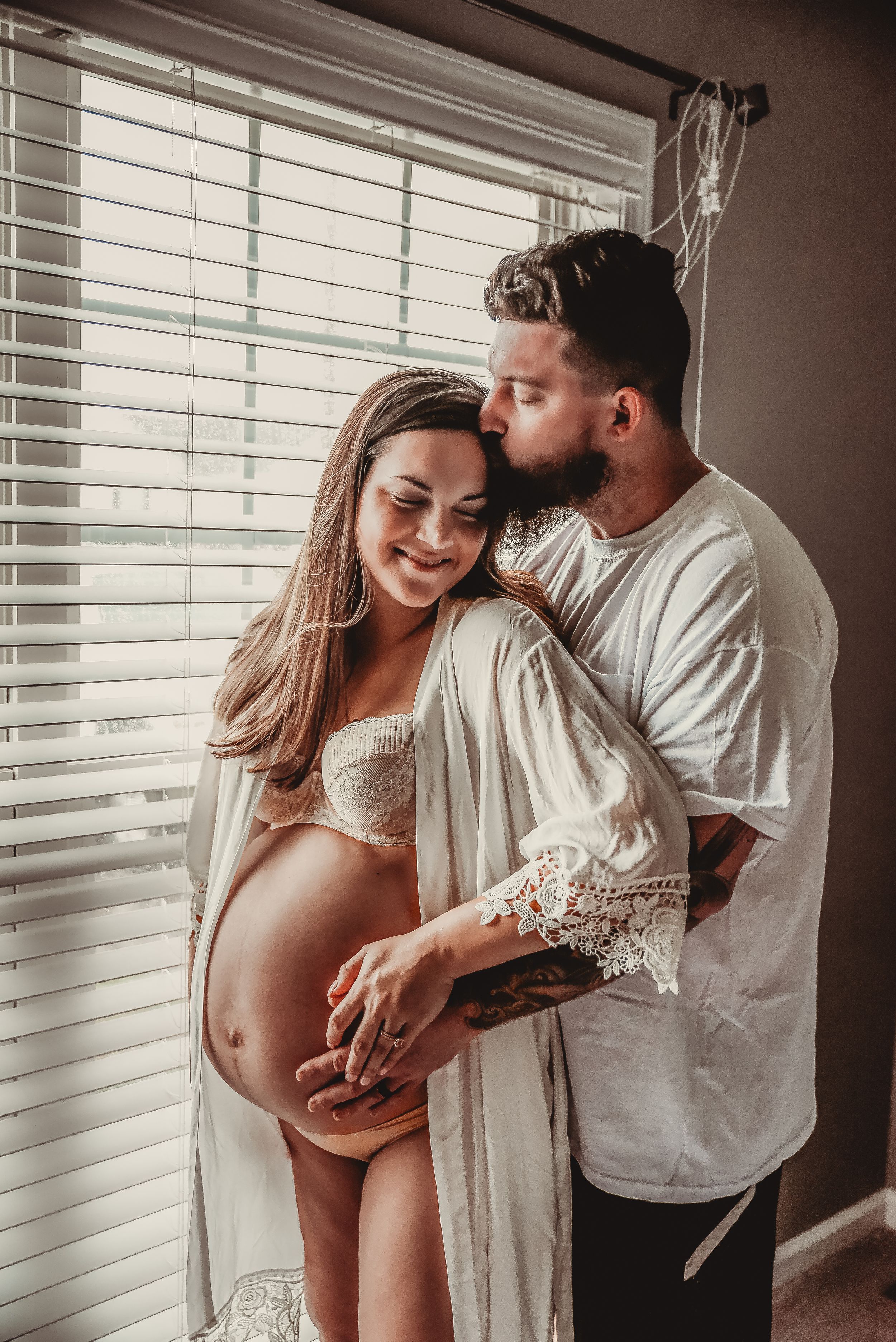 Couple sharing tender moments during maternity photoshoot near window with venetian blinds.