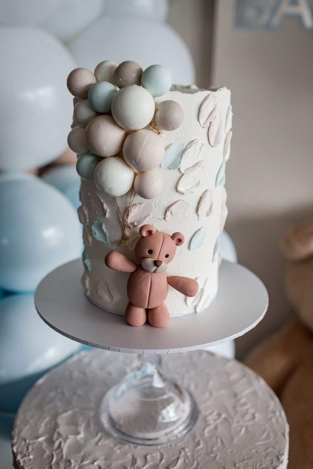 A white baby shower cake decorated with pastel balloons and a teddy bear topper.