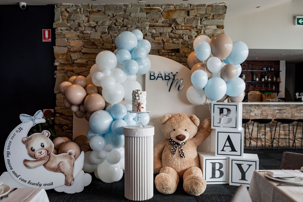 Baby shower decoration setup with balloon arch, teddy bear, and white blocks against stone wall.