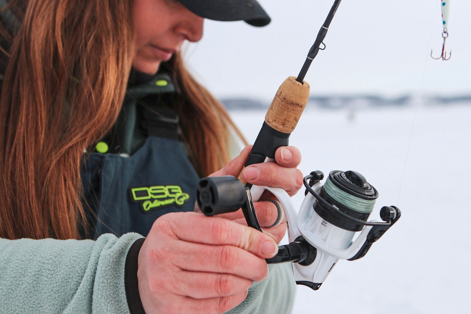 Close up of hands holding an ice fishing rod with a white spinning reel against a snowy background.
