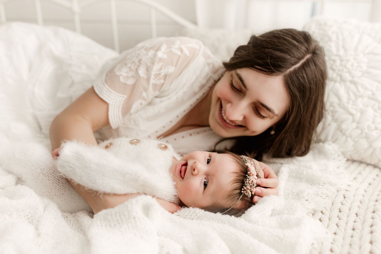 Smiling brunette mother lies on white knit blanket with adorable baby wearing floral headband.