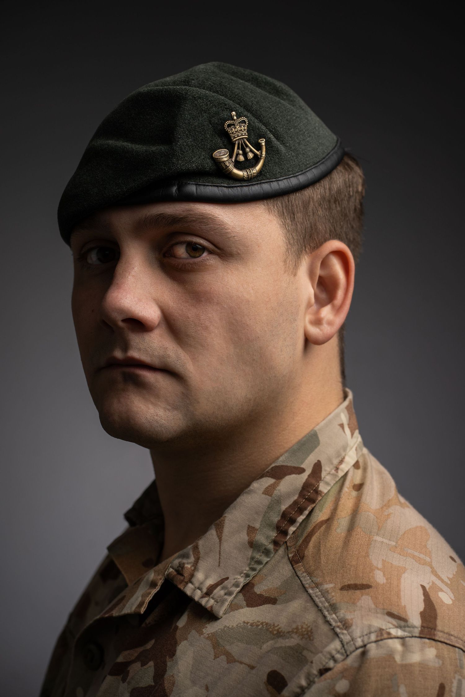 Military service member in desert camouflage uniform and green beret against gray background.