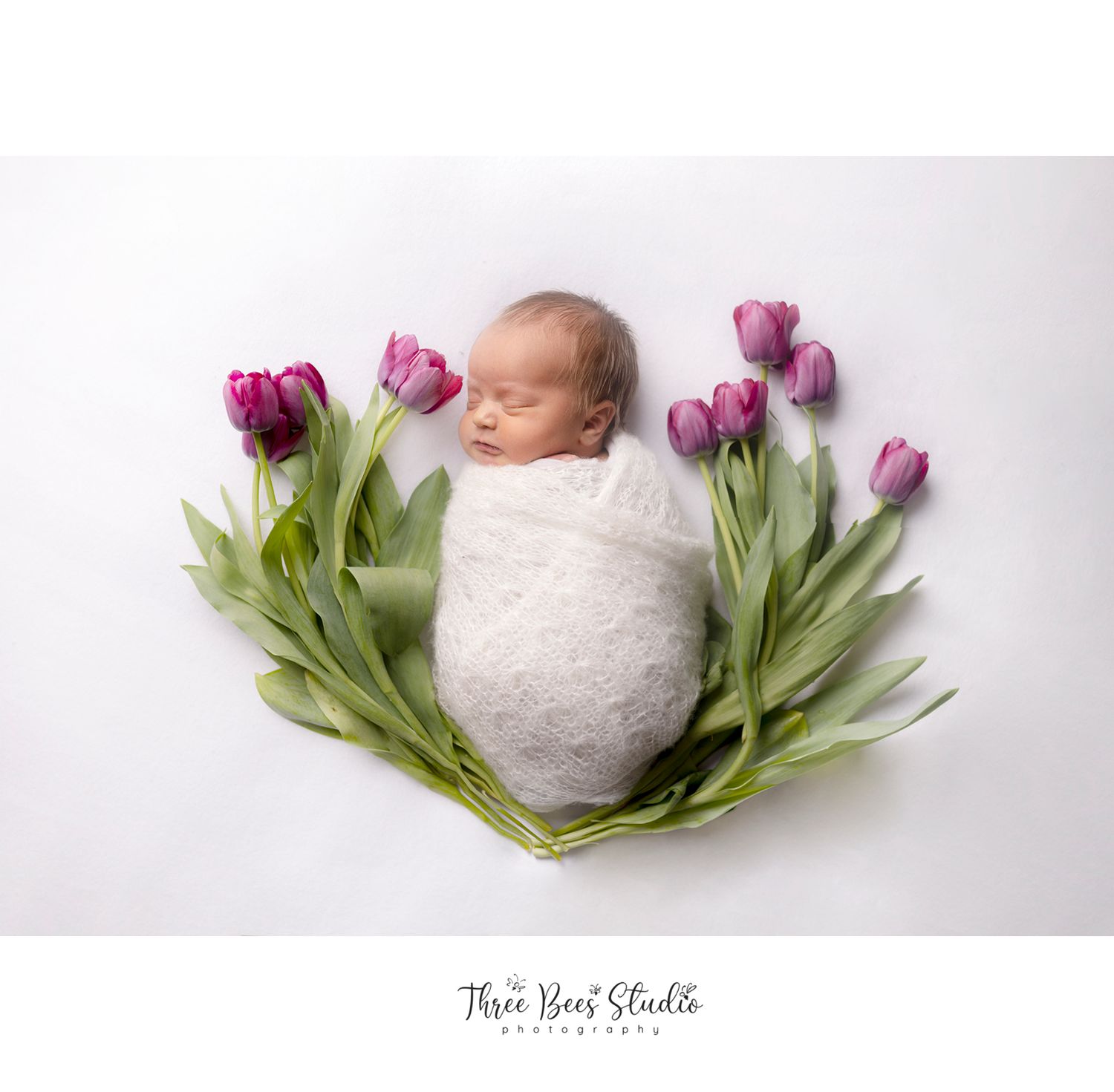Newborn wrapped in white swaddle surrounded by vibrant purple tulips on white background.