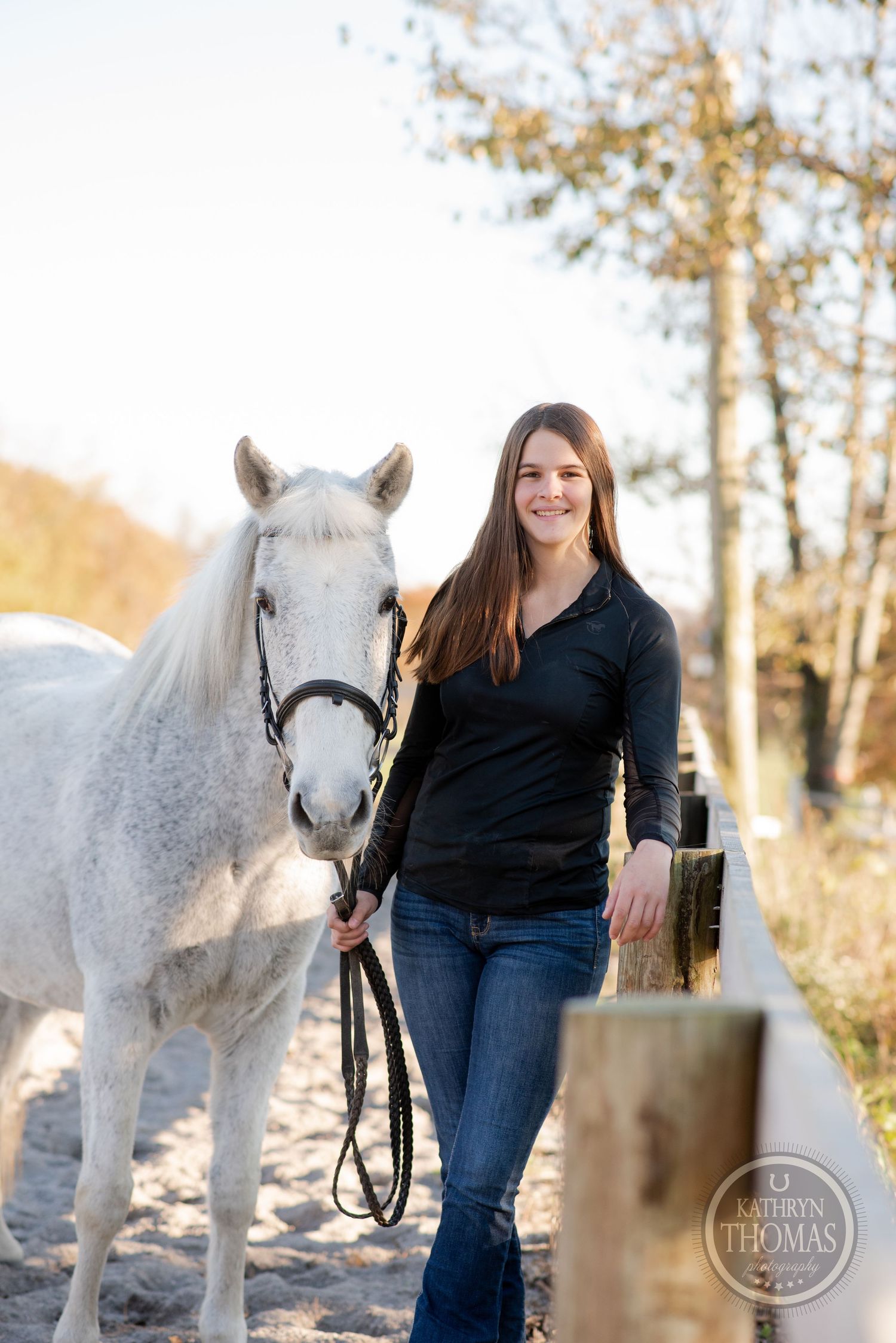 A beautiful white horse stands with a companion in casual attire against autumn trees.
