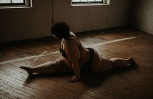 A person stretches in a split position on a wooden floor in soft natural light from nearby windows.