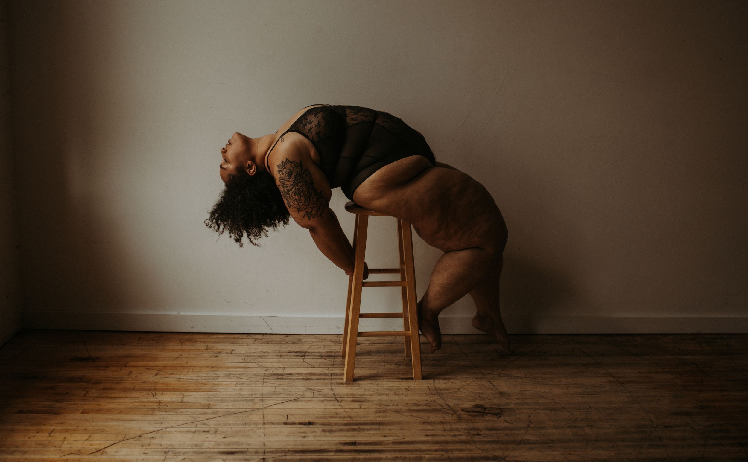 Moody boudoir portrait on wooden stool with dramatic shadowing and minimal styling.