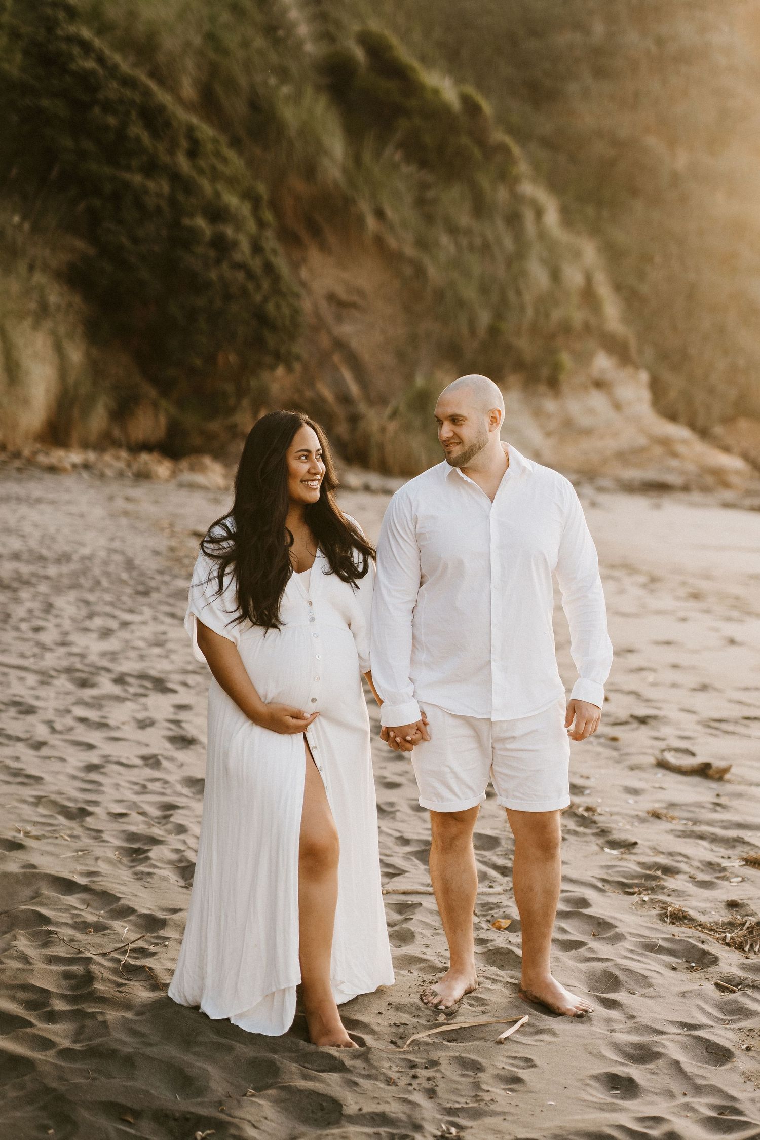 Expecting couple in white outfits hold hands during sunset beach maternity photoshoot.