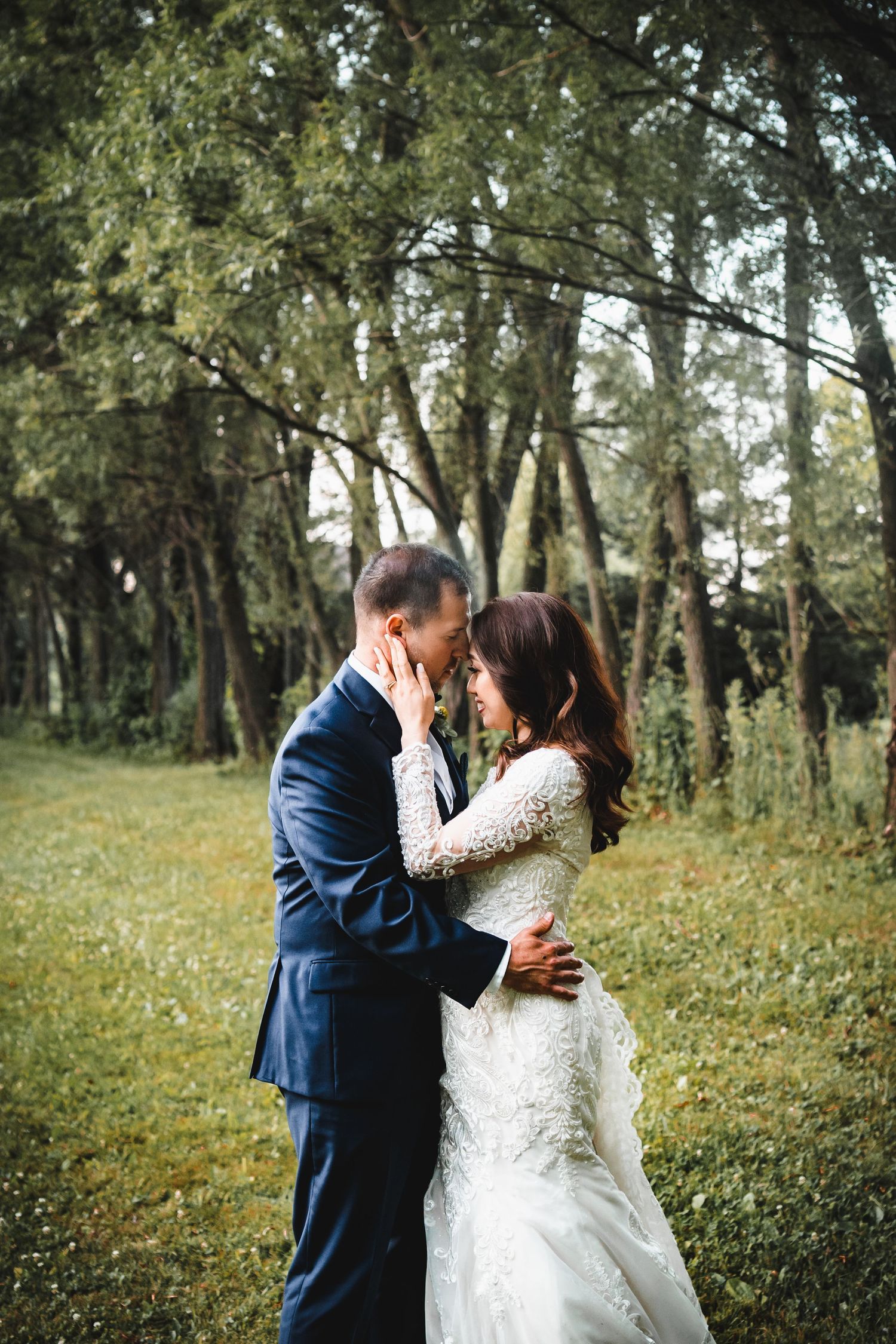 Romantic wedding couple embracing in sunlit wooded area with bride in lace gown.