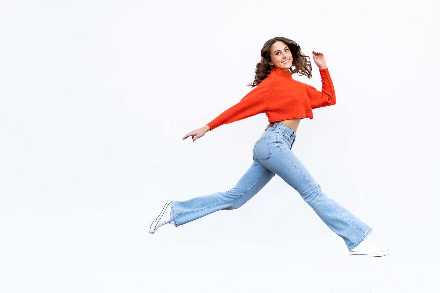 A high school senior in red sweater and flared jeans jumping joyfully against white background.