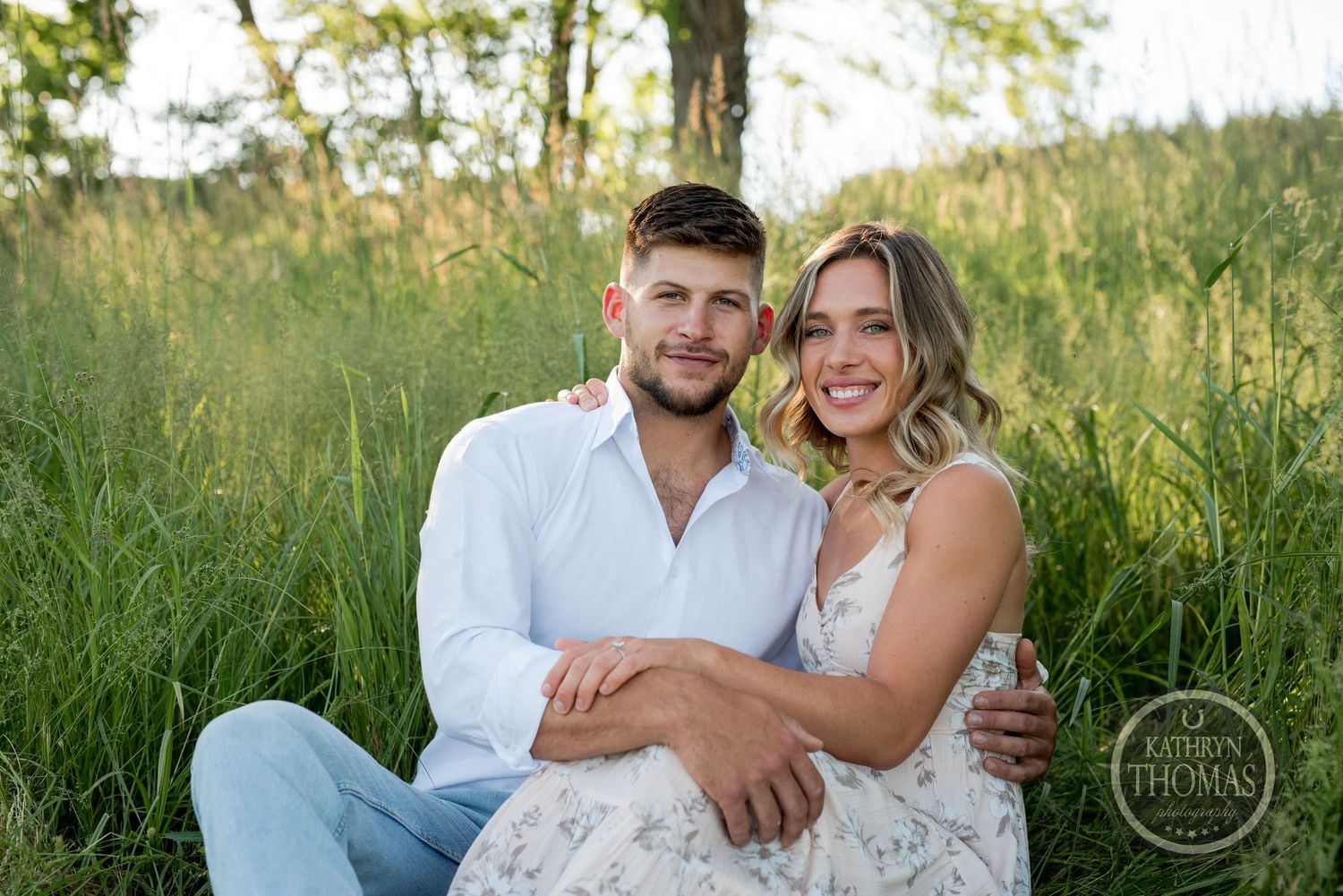 A happy couple sits together in tall grass during a summer photo session.
