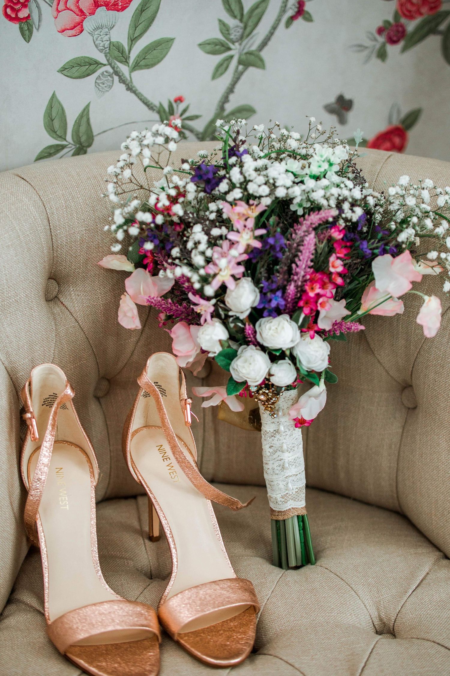 Wedding accessories feature rose gold heels and a vibrant wildflower bouquet on a beige tufted chair.