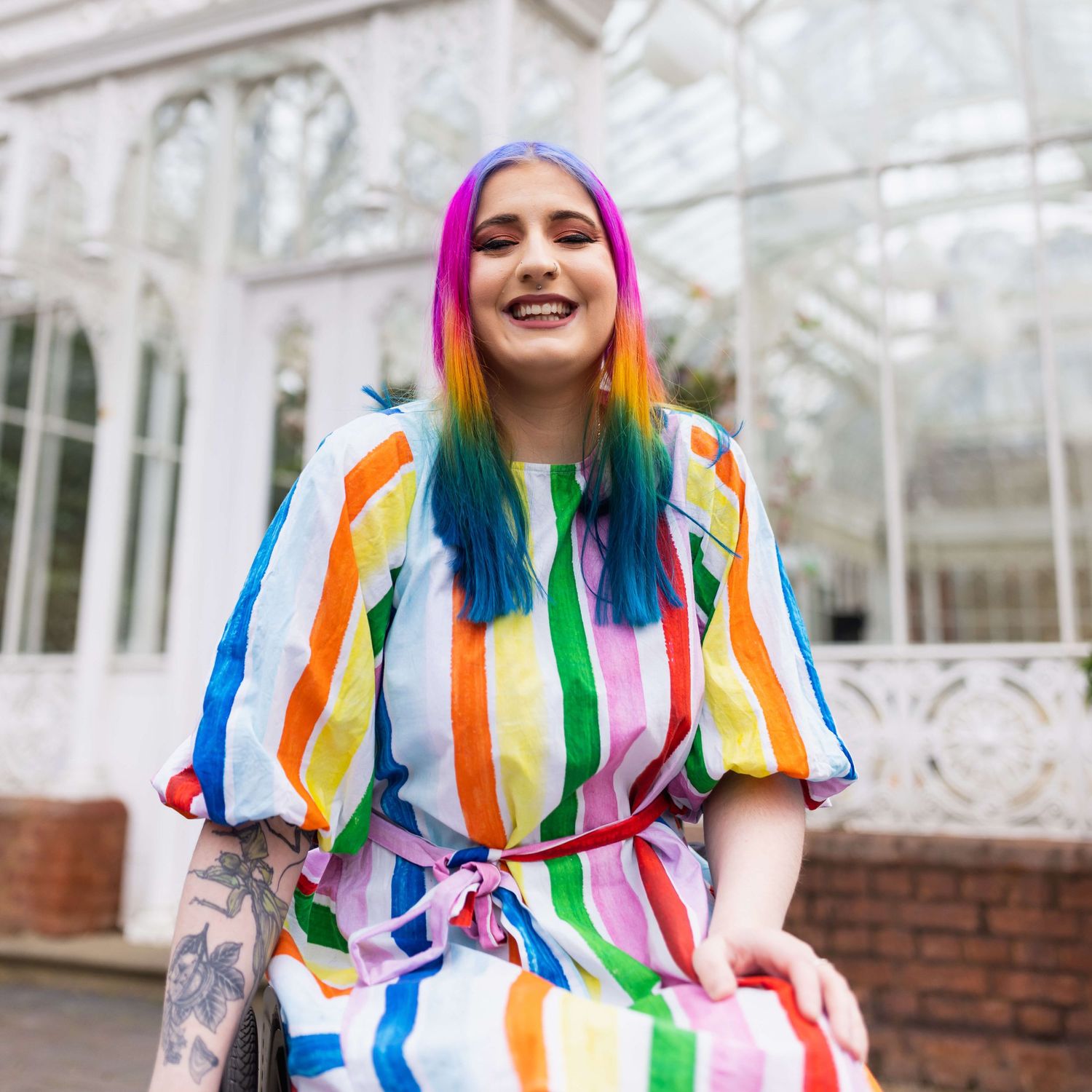 London rainbow haired disabled influencer posing sat smiling in their wheelchair wearing a rainbow outfit.