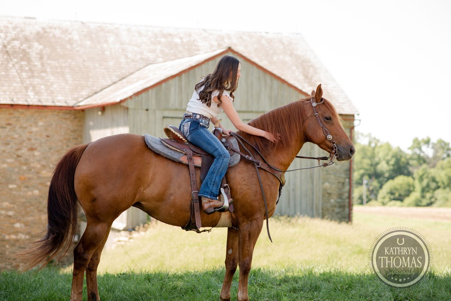 A person rides a brown horse in front of a rustic barn during a summer day.