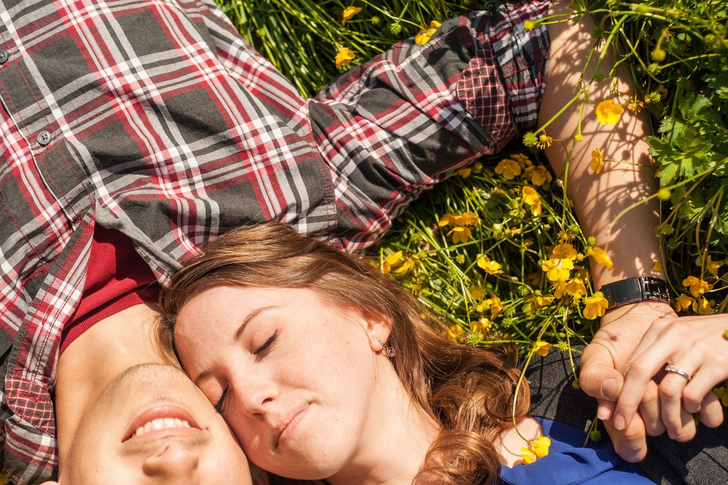 Couple lying in grass surrounded by yellow flowers on sunny day.
