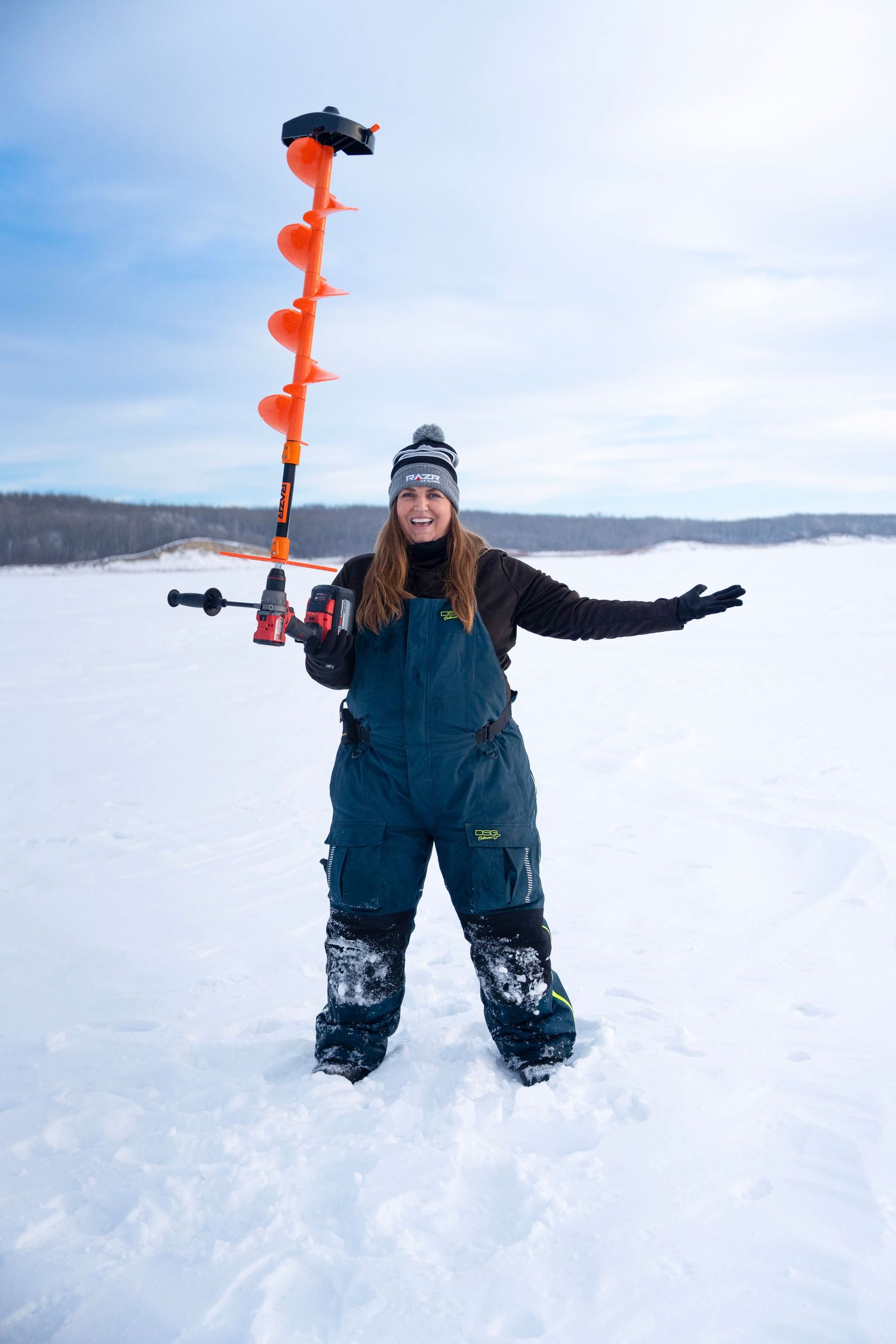 Orange ice auger drill held up in snowy winter landscape during ice fishing.