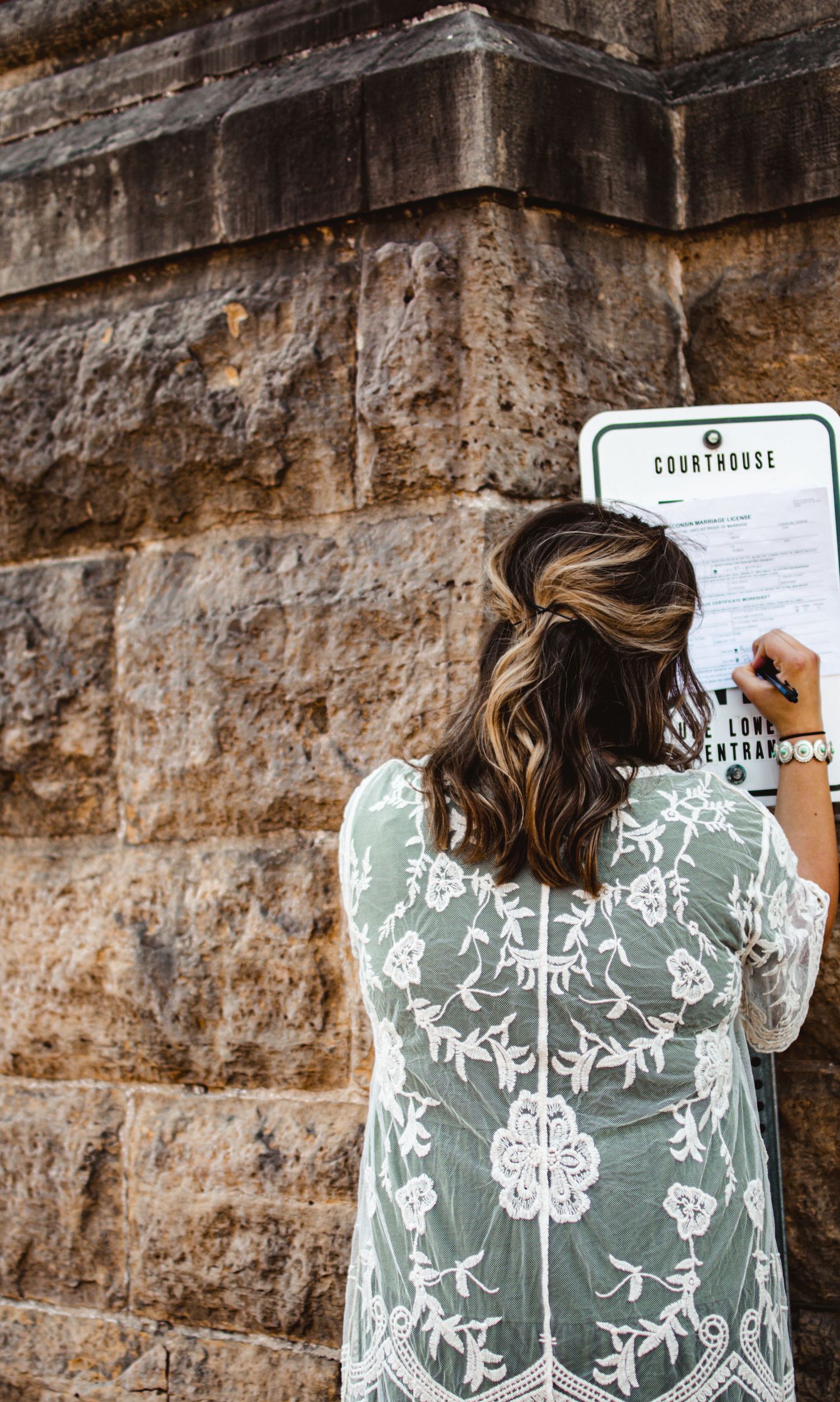 Person in embroidered dress writing on clipboard against stone wall.
