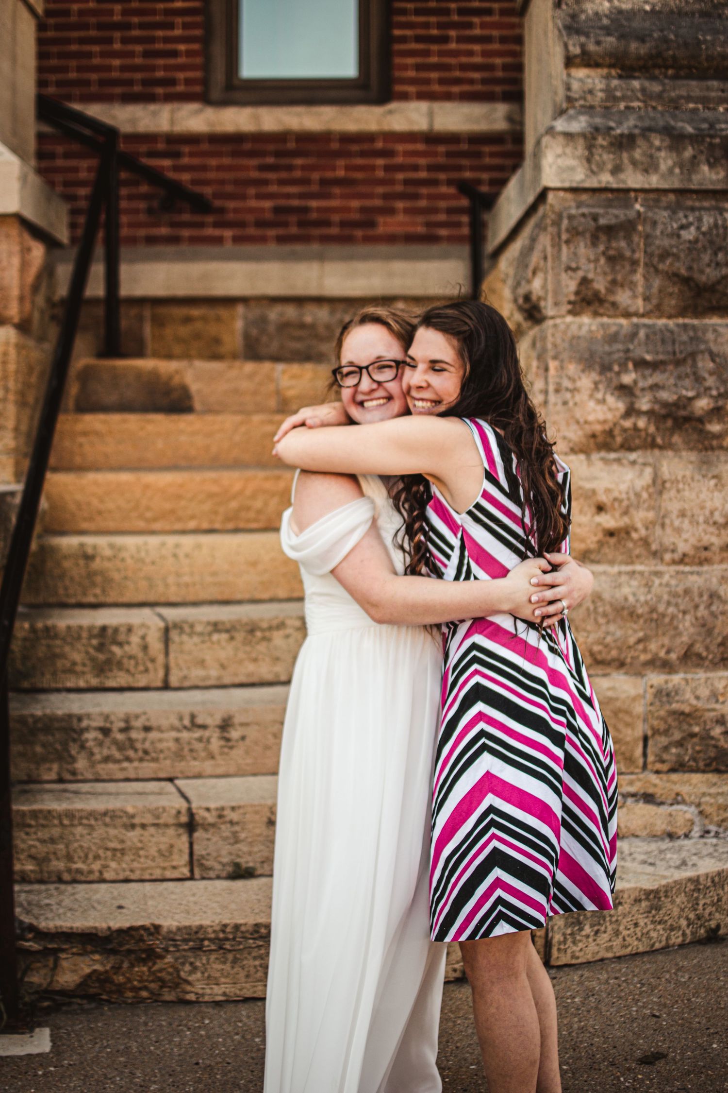 Two people in colorful dresses share a warm embrace on stone steps.