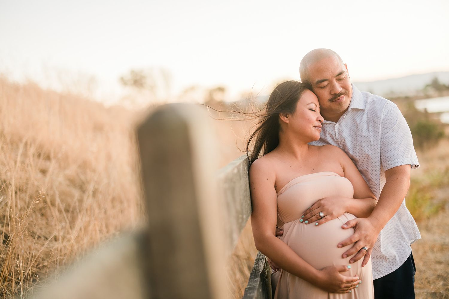 Intimate maternity photo session captured at sunset with a fence in soft lighting.
