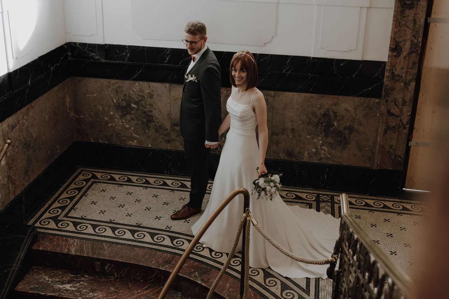 Couple stands on ornate staircase with vintage marble and tile details.