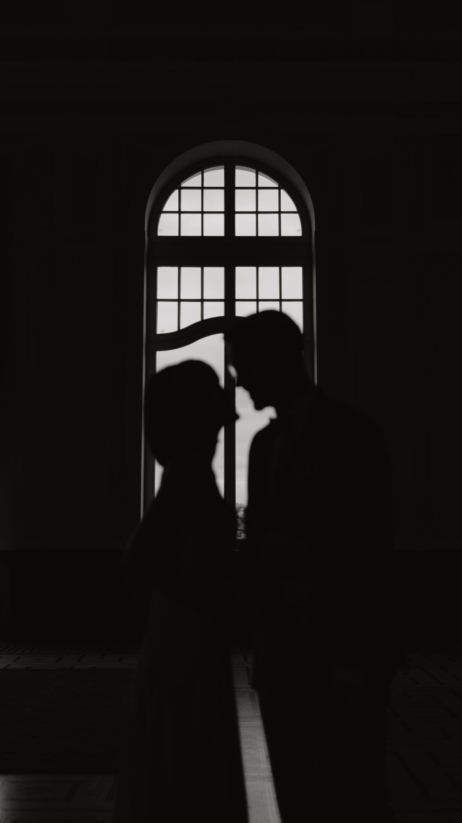 Silhouetted couple embracing in front of arched window creating dramatic contrast.