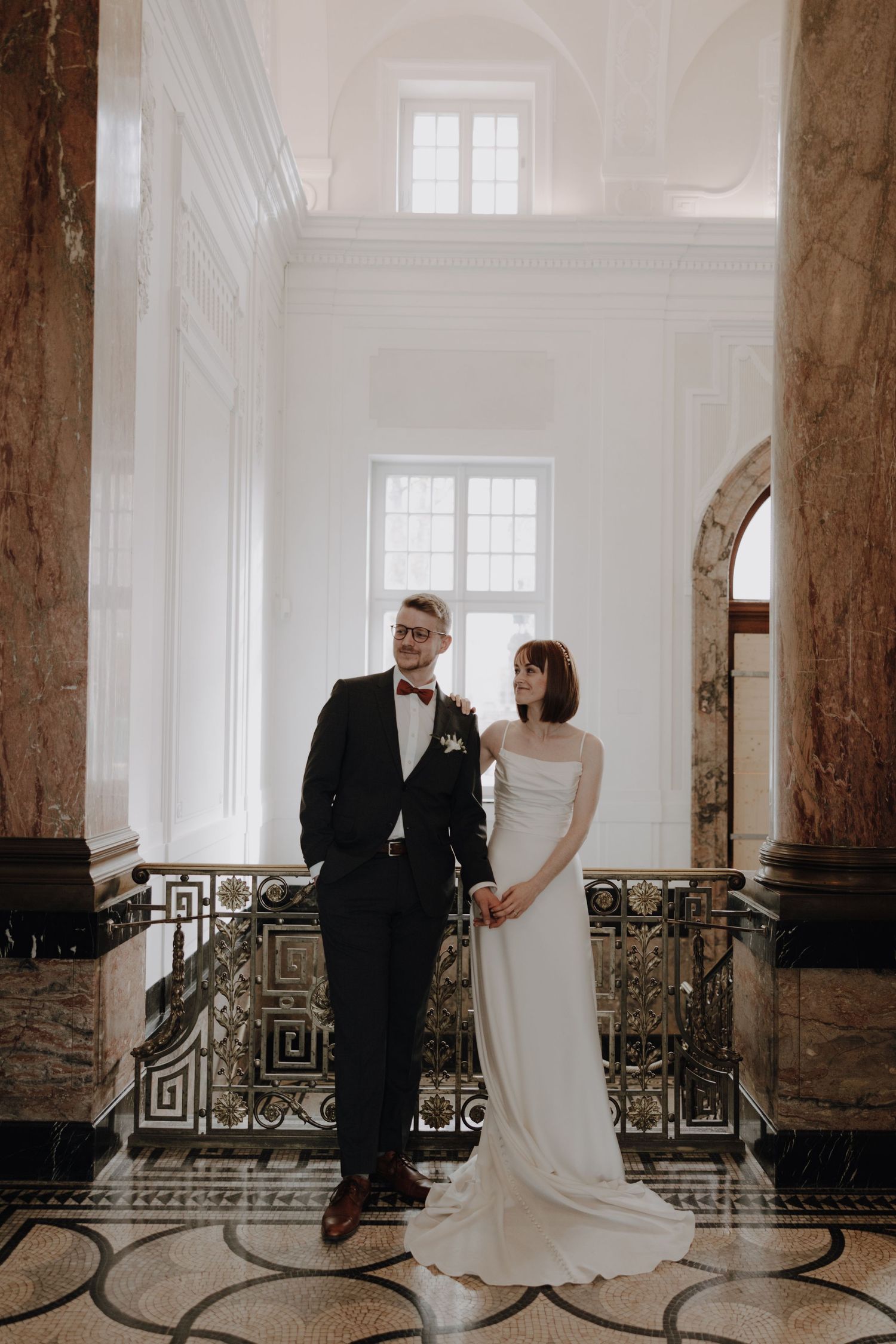 An elegant couple shares an intimate moment in a bright hallway with classical stone columns.