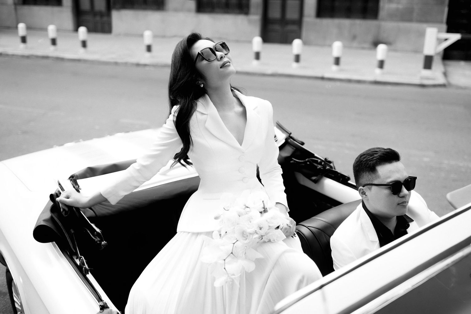 A glamorous black and white photo of a couple posing with a vintage convertible car on a city street.