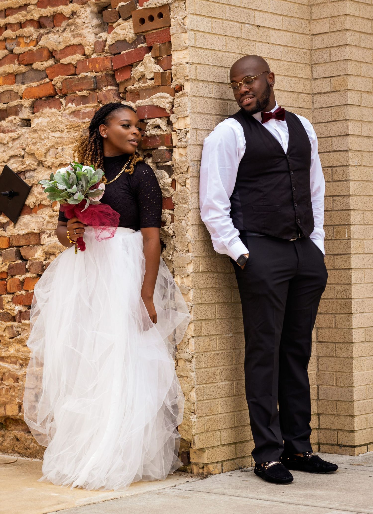 Wedding couple poses against brick wall, showcasing elegant attire and floral bouquet.