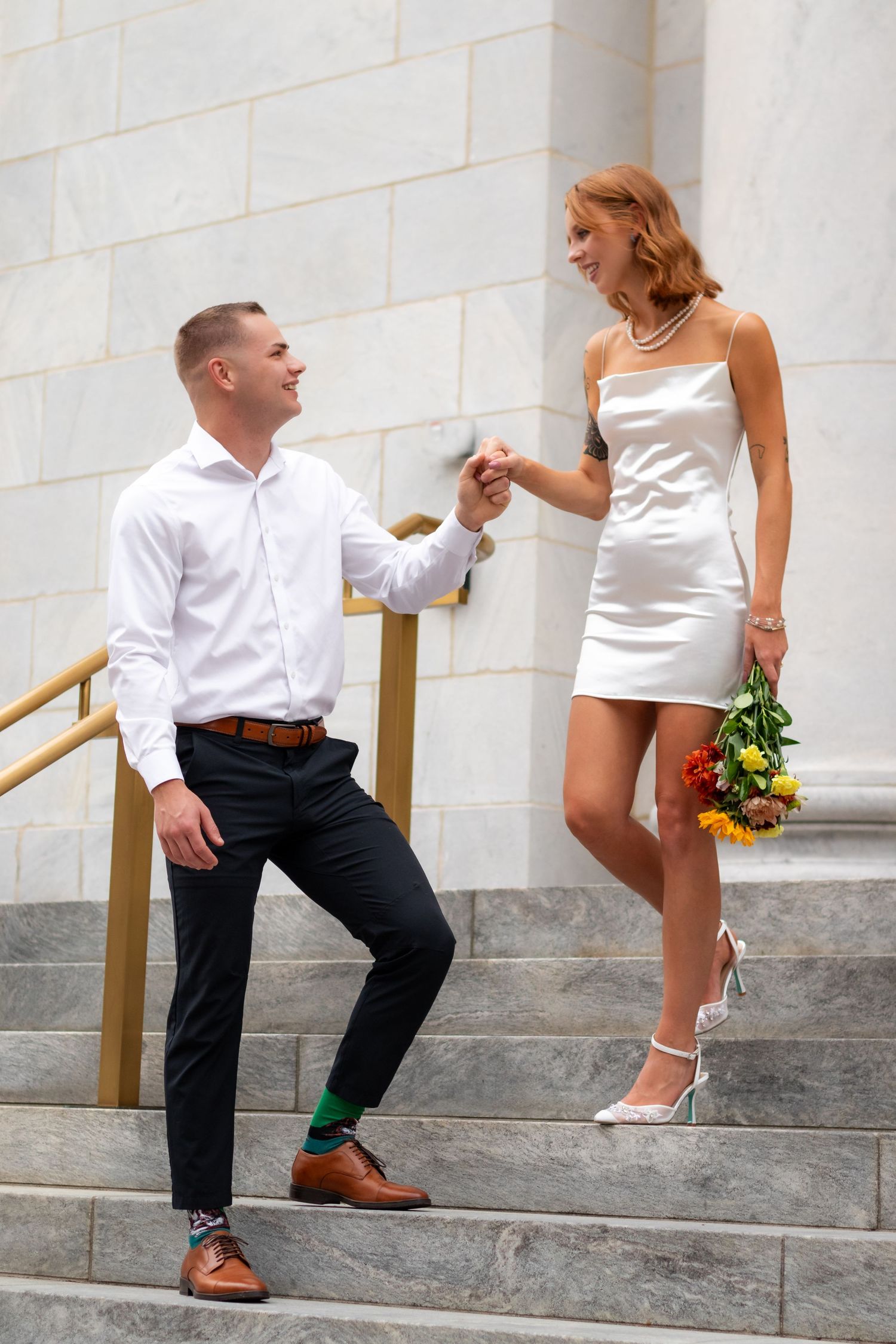 Playful couple poses on stone steps with bride in short white dress.