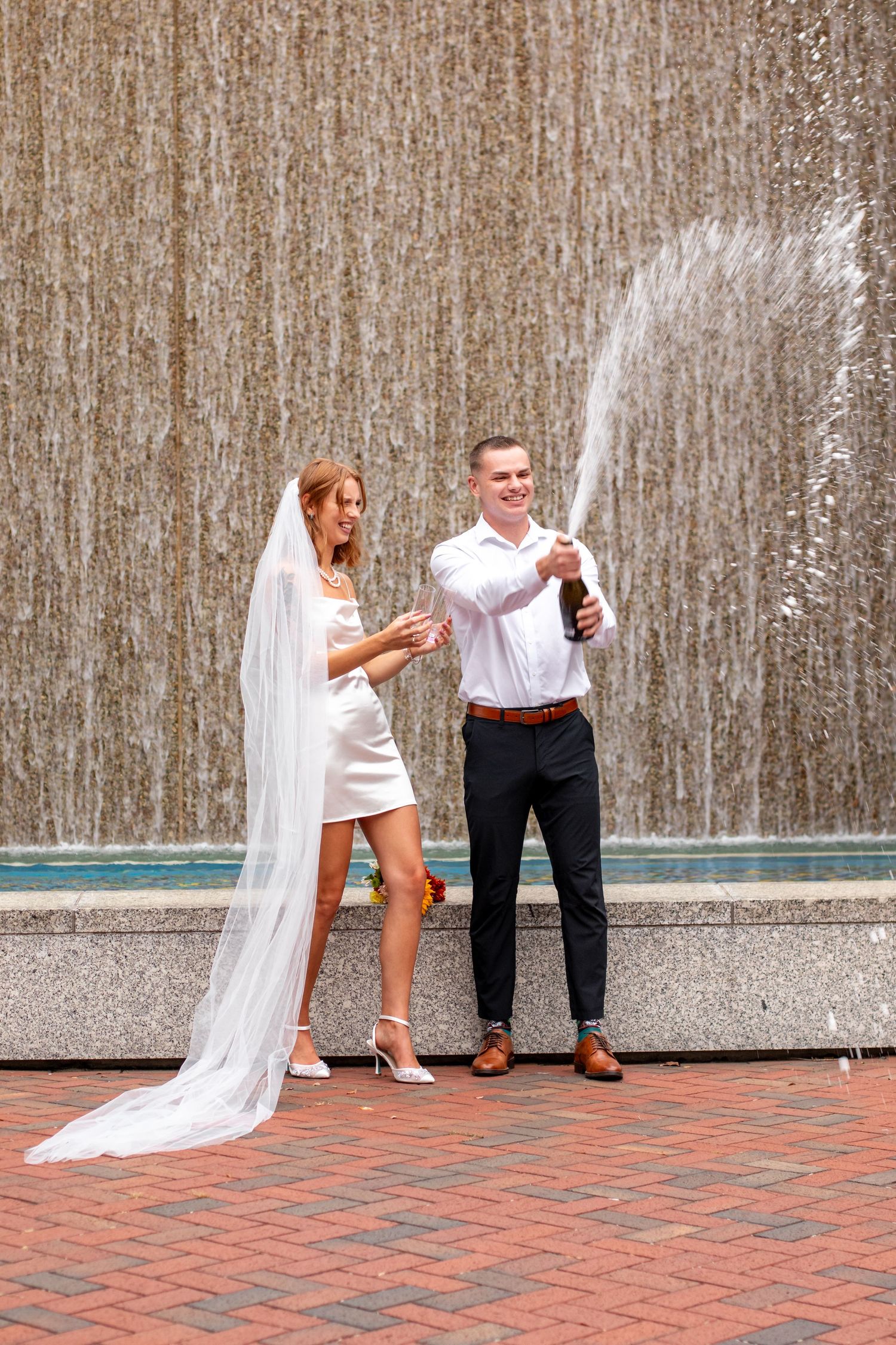 A couple pops champagne in front of a waterfall wall, with her wearing a white dress and veil.