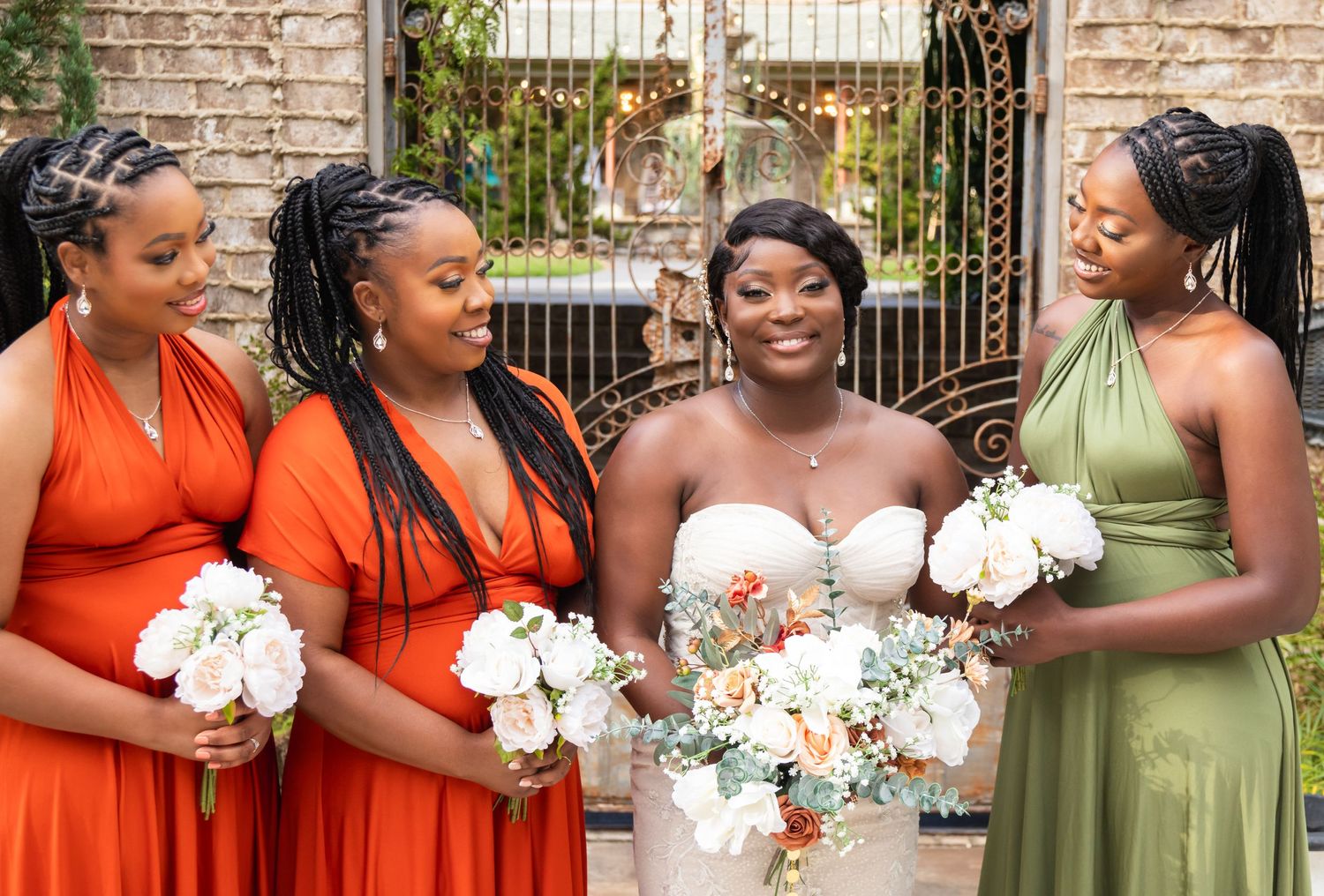 Bridesmaids wearing orange and green dresses hold white bouquets while standing alongside the bride at a rustic venue.