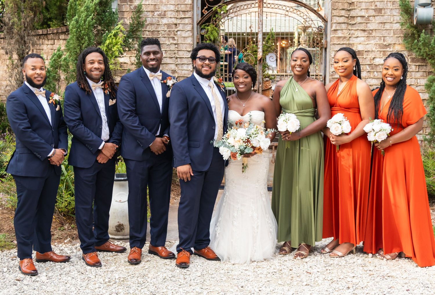 Wedding party in navy suits and orange and green dresses pose together in front of a brick wall.