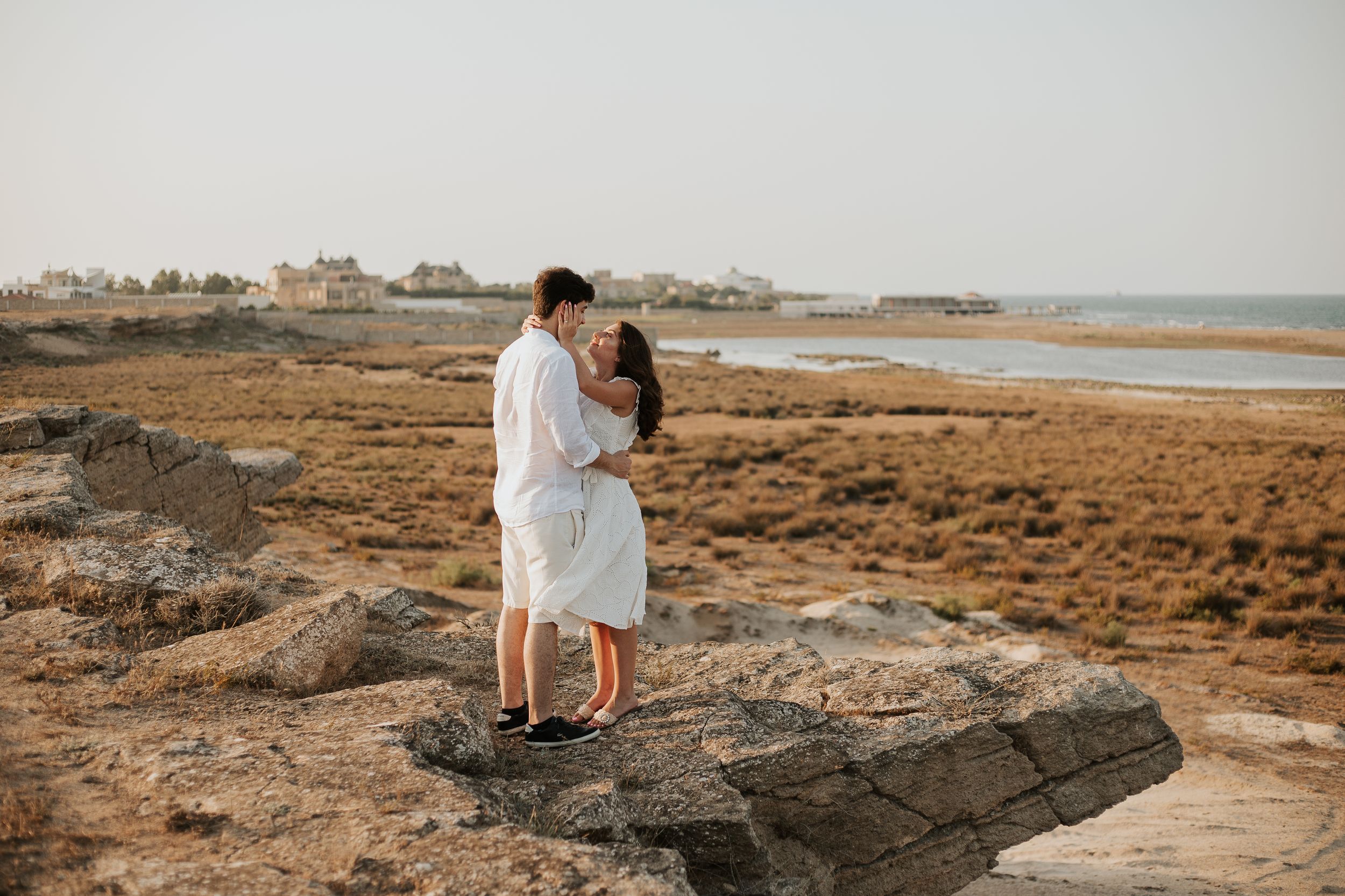 A couple embraces on coastal rocks with ocean and buildings visible in background.