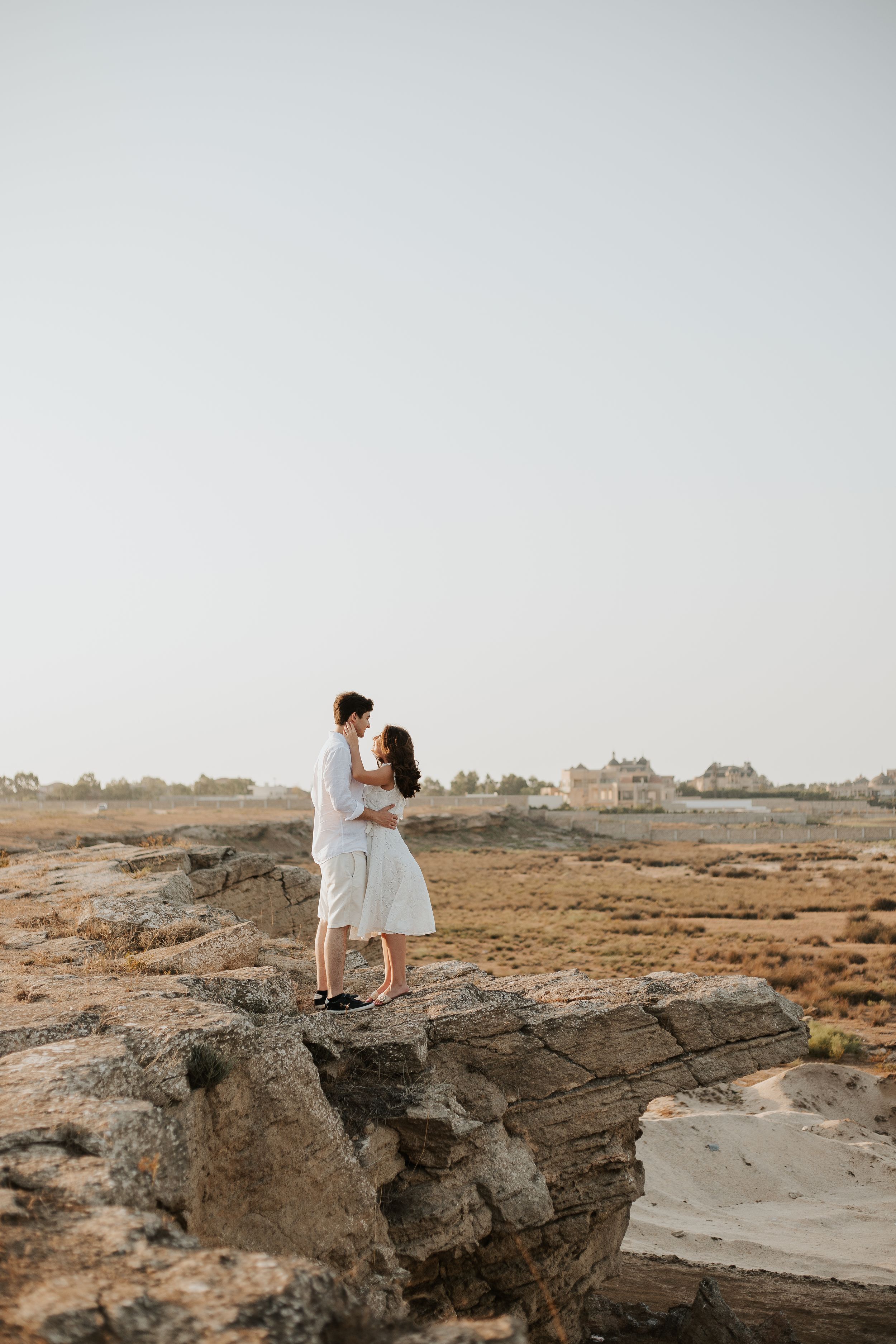 A couple stands on rocky cliffs overlooking a desert landscape at sunset.