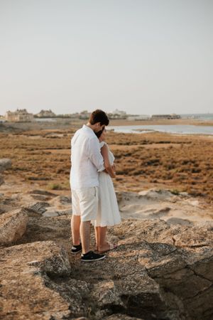 A couple in white outfits embraces while standing on rocky coastal terrain at sunset.