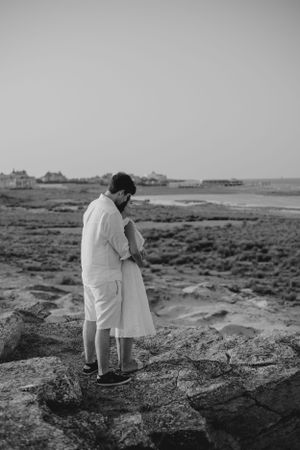 Black and white photo of a couple embracing on rocky coastal terrain with houses in distance.