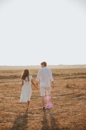 Couple dressed in white holds hands while walking through an open field at golden hour.