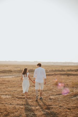 Two people walking hand in hand across a golden sunlit field wearing coordinated white outfits.