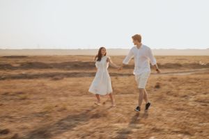 A couple in white outfits joyfully walks through a sun-drenched field.