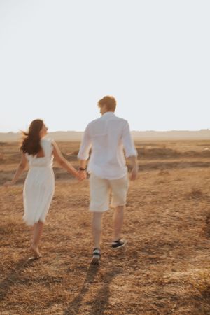 Two people in white clothing walk hand in hand across a golden sunset landscape.