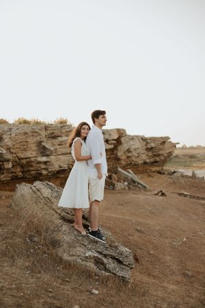Couple in white outfits standing on rocky cliff edge at sunset.